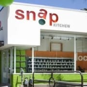 A modern storefront with large windows and the sign "snap KITCHEN" in red and green letters above the entrance, this spot stands out among healthy Austin restaurants. The building exterior is white with green accents. Austin Marathon Half Marathon & 5K