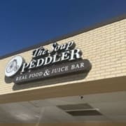 A sign for "The Soup Peddler Real Food & Juice Bar," one of the top Austin Healthy Dining spots, is mounted on a light brick building under a clear blue sky. The sign features a bicycle graphic on the left. Austin Marathon Half Marathon & 5K