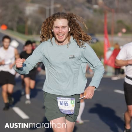 A smiling man with long curly hair, wearing a light green shirt and shorts, runs in a marathon with a race bib numbered 6755, burning calories as he goes. Other runners are visible in the background on this sunny day. Austin Marathon Half Marathon & 5K