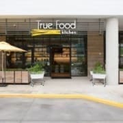 Exterior view of True Food Kitchen, one of the top Healthy Austin Restaurants, featuring a modern entrance with large windows, potted plants, and a yellow umbrella shading outdoor seating. The restaurant sign is visible above the door. Austin Marathon Half Marathon & 5K