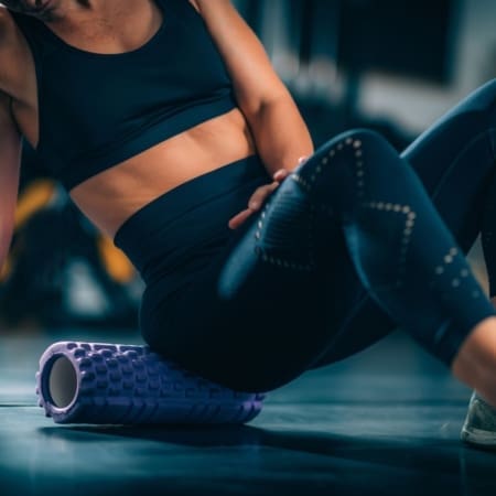A person in black athletic wear practices foam rolling on the gym floor to massage their lower back and hip muscles—an essential recovery routine for Marathon Success. The focus is on the torso and legs, with gym equipment blurred in the background. Austin Marathon Half Marathon & 5K