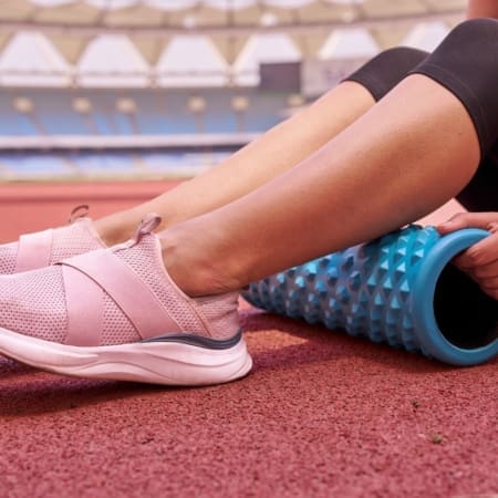 A person wearing pink athletic shoes uses a textured blue foam roller for foam rolling their calf while sitting on a running track, possibly training for the Austin Marathon in a stadium. Austin Marathon Half Marathon & 5K
