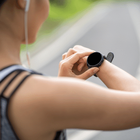 A person wearing sportswear and earphones stands outside on a paved path, checking a black smartwatch on their wrist. The uplifting scene hints at running for better mental health, with a blurred roadway and greenery in the background. Austin Marathon Half Marathon & 5K
