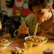 A young man uses chopsticks to eat noodles from a bowl at a wooden table, with other food dishes and a drink nearby—an inviting scene that could inspire any runner’s diet. Warm sunlight brightens the meal, highlighting balanced nutrition. Austin Marathon Half Marathon & 5K