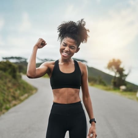 A young woman in athletic wear stands on a road, smiling and raising her fist in triumph. She radiates marathon motivation and happiness, with greenery and a bright sky in the background. Austin Marathon Half Marathon & 5K
