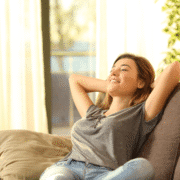 A young woman enjoys a well-deserved rest on a couch with her hands behind her head, smiling and looking content. Sunlight streams through large windows in the background, creating a warm, peaceful atmosphere—perfect for recovery days during marathon training. Austin Marathon Half Marathon & 5K
