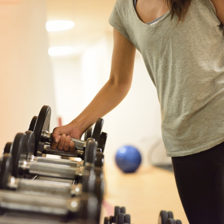 A person wearing a gray t-shirt and black pants picks up a dumbbell from a rack in a gym, focusing on strength training for runners. Exercise equipment and a blurred blue yoga ball are visible in the background. Austin Marathon Half Marathon & 5K