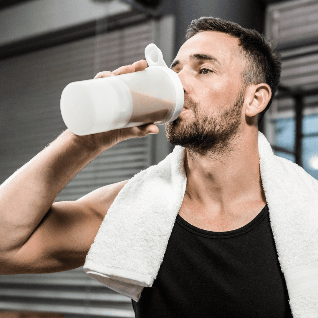 A man with a beard and short hair, wearing a black tank top and a white towel around his neck, drinks from a protein shaker bottle in a gym setting—highlighting what runners should eat post-workout. Austin Marathon Half Marathon & 5K