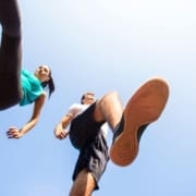 Two people, a woman and a man, are seen from below as they walk or jog outdoors. The bright blue sky is in the background, and one man’s sneakered foot is prominently in the foreground, showcasing proper running form. Austin Marathon Half Marathon & 5K
