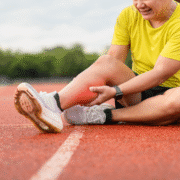 A person in a yellow shirt sits on a running track, holding their lower leg with a pained expression—possibly from marathon training. The affected area appears slightly red, highlighting the importance of rest days for proper recovery. Austin Marathon Half Marathon & 5K