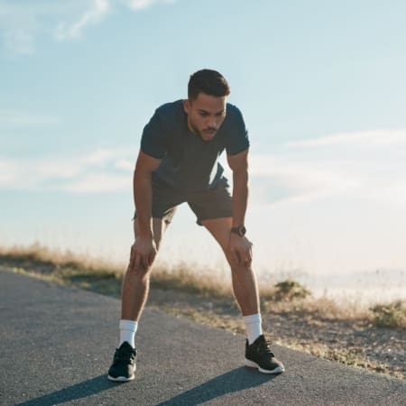 A man in athletic wear rests with his hands on his knees on an outdoor path, appearing tired after marathon training, with a clear sky and nature in the background. Austin Marathon Half Marathon & 5K
