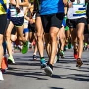 A group of runners in athletic gear and numbered bibs participate in a road race, focusing on their running form and making small adjustments for big gains as they move along a paved street under bright sunlight. Austin Marathon Half Marathon & 5K
