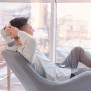 A man sits in a modern chair by a large window, leaning back with his hands behind his head and looking relaxed. Soft daylight fills the room, reflecting the calm and peaceful atmosphere of well-earned rest on recovery days from marathon training. Austin Marathon Half Marathon & 5K