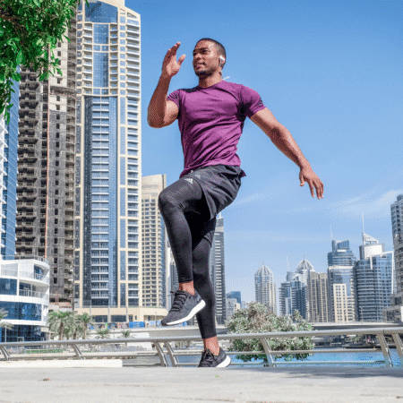 A man in athletic wear jogs in place outdoors on a sunny day, with tall modern buildings and blue sky in the background, focusing on building power and strength training for runners without bulking up. Austin Marathon Half Marathon & 5K