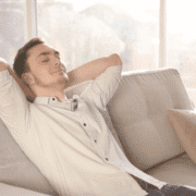 A young man with short hair and a light beard is relaxing on a beige sofa with his hands behind his head, eyes closed in calm rest. Sunlight streams through large windows—a perfect recovery day after marathon training. Austin Marathon Half Marathon & 5K