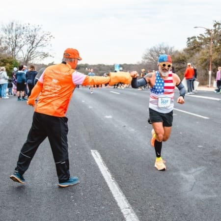 A runner in a USA-themed tank top and blue headband, fueled by marathon motivation, fist bumps a volunteer in orange during a road race, with spectators and other runners cheering in the background. Austin Marathon Half Marathon & 5K