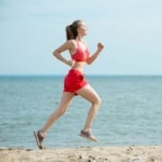 A woman in red athletic wear jogs along a sandy beach, showcasing excellent running form as the ocean sparkles in the background on a clear day. Austin Marathon Half Marathon & 5K