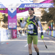 A smiling runner in a green and black athletic outfit and neon gloves celebrates while crossing the Austin Marathon finish line at his ideal marathon pace, holding a cane in one hand. Colorful banners and cheering spectators fill the background. Austin Marathon Half Marathon & 5K
