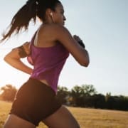 A woman in athletic wear runs outdoors in a park at sunrise or sunset, listening to music with earphones. The sky is clear and trees are visible in the background, as she focuses on her running form. Austin Marathon Half Marathon & 5K