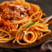 A close-up of spaghetti with meat sauce on a dark plate, topped with grated cheese and a basil leaf. A fork is twirling some spaghetti in the foreground, showcasing a classic example of what runners eat for balanced runners nutrition. Austin Marathon Half Marathon & 5K