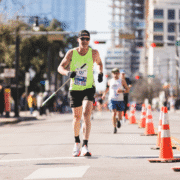 A male runner in sunglasses and a neon green tank top runs on a city street during a race, smiling as he maintains his ideal marathon pace, with traffic cones marking the route and other runners visible in the background. Austin Marathon Half Marathon & 5K