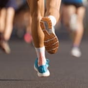Close-up of a runner’s legs and shoes from behind, mid-stride on a road during a race, highlighting strong running form. The runner wears colorful socks and blue athletic shoes with orange soles, with other runners blurred in the background. Austin Marathon Half Marathon & 5K