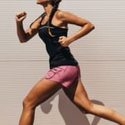 A woman in a black tank top and pink shorts is running outdoors against a light-colored, ridged wall. Her hair is in braids, and she appears to be mid-stride, showcasing excellent running form as she focuses on her exercise. Austin Marathon Half Marathon & 5K