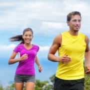 A man in a yellow sleeveless shirt and a woman in a purple shirt are jogging outdoors with proper running form, smiling, under a blue sky with greenery in the background. Austin Marathon Half Marathon & 5K