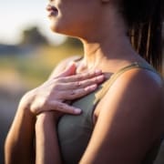 A woman stands outdoors with both hands placed gently over her chest, wearing a sleeveless top. The softly blurred background suggests a calm setting, perfect for reflecting on running improvements or refining her running form. Austin Marathon Half Marathon & 5K