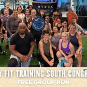 A diverse group of people in workout clothes pose and smile together inside a gym, holding a Body Fit Training sign. The text overlay reads: "Body Fit Training South Congress free group run. Austin Marathon Half Marathon & 5K