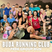 A diverse group of smiling people poses together outdoors before a Buda Running Club group run, with some making playful gestures. Text below reads: "Buda Running Club Free Group Run" on a decorated banner. Austin Marathon Half Marathon & 5K