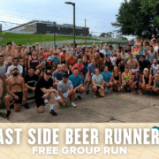 A large group of people pose outdoors for a group photo before a free group run, with greenery and a building in the background. The text overlay reads: "East Side Beer Runners Free Group Run. Austin Marathon Half Marathon & 5K