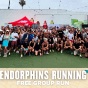 A large, diverse group of people in sporty attire smile and pose outdoors in front of surfboards and shops. A banner at the bottom reads "Endorphins Running Free Group Run," celebrating their energizing group run. Austin Marathon Half Marathon & 5K
