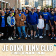 A large group of smiling people, some in blue "VOLUNTEER" shirts, pose outdoors in front of city buildings for a photo at the JE DUNN RUNN CLUB free run event. A dog is also in the front row. Austin Marathon Half Marathon & 5K