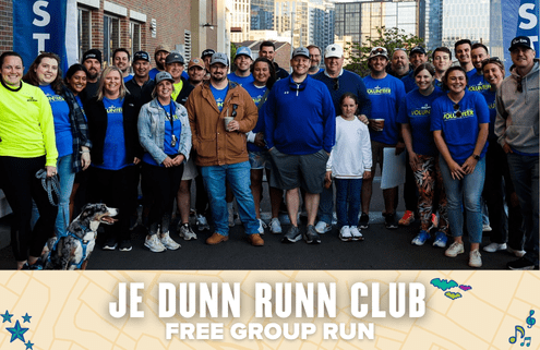 A large group of smiling people, some in blue "VOLUNTEER" shirts, pose outdoors in front of city buildings for a photo at the JE DUNN RUNN CLUB free run event. A dog is also in the front row. Austin Marathon Half Marathon & 5K