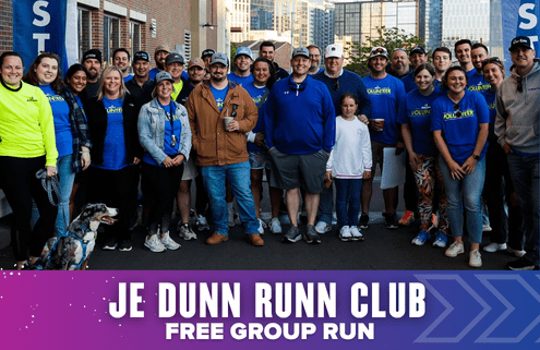 A diverse group of people, some in "VOLUNTEER" shirts, smile and pose outside for a photo with a dog in front. Text reads: "JE Dunn Running Club Free Group Run." City buildings rise in the background. Austin Marathon Half Marathon & 5K