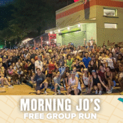 A large, diverse group of people pose together outside a brightly lit building at night. A banner at the bottom reads, “Morning Jo’s Free Group Run,” with a map and playful icons as the background. Join Morning Jo's for a free run!. Austin Marathon Half Marathon & 5K