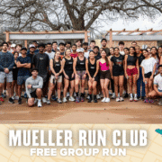 A large, diverse group of people in athletic wear pose and smile outdoors for a group photo. Text at the bottom reads: "Mueller Run Club - Free Group Run." Trees and wooden benches frame this lively free run gathering. Austin Marathon Half Marathon & 5K