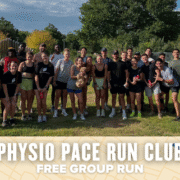 A large group of smiling people in athletic wear pose together outdoors on grass at the Physio Pace group run, some holding water bottles or a small dog. The banner reads "Physio Pace Run Club Free Group Run," with trees and buildings in the background. Austin Marathon Half Marathon & 5K