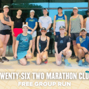 A group of 14 runners, dressed in athletic gear and sneakers, pose and smile together outside a building. Text at the bottom reads: "Twenty-Six Two Marathon Club Free Group Run" with decorative icons, highlighting this free group run event. Austin Marathon Half Marathon & 5K