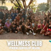 A large, diverse group of people pose and smile together outdoors after a Wellmess Club free group run, with a small dog at the front. Text at the bottom reads, "WELLMESS CLUB FREE GROUP RUN. Austin Marathon Half Marathon & 5K