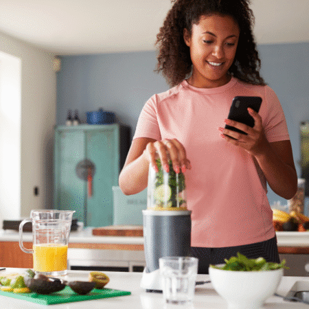 A woman in a pink shirt uses a blender with greens and lemon slices while looking at her smartphone in a bright kitchen, possibly tracking progress. A pitcher of orange juice and chopped avocado are on the counter. Austin Marathon Half Marathon & 5K