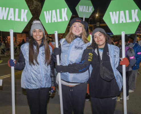 Three women stand side by side at night, each holding a green octagonal "WALK" sign. Dressed warmly and smiling, they embrace volunteer opportunities, with other people and city lights glowing in the background. Austin Marathon Half Marathon & 5K