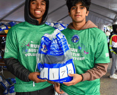 Two young men wearing green volunteer shirts hold a clear bag labeled "1170" under a tent at an outdoor event, smiling at the camera. The scene highlights volunteer opportunities, with others and bags visible in the background. Austin Marathon Half Marathon & 5K