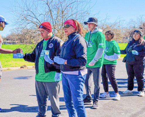 A group of people in casual clothes and gloves stand outdoors on a sunny day, handing out cups at a community event, showcasing local volunteer opportunities. Leafless trees and green grass are visible in the background. Austin Marathon Half Marathon & 5K