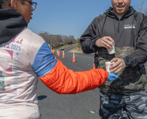 A volunteer explores new volunteer opportunities by handing a cup of water to a runner during a race on a sunny day, with orange cones and a clear blue sky in the background. Austin Marathon Half Marathon & 5K
