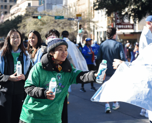 A woman in a green shirt and winter hat hands out water bottles to runners during an outdoor event on a city street, highlighting volunteer opportunities amid a team of helpers and tall buildings in the background. Austin Marathon Half Marathon & 5K