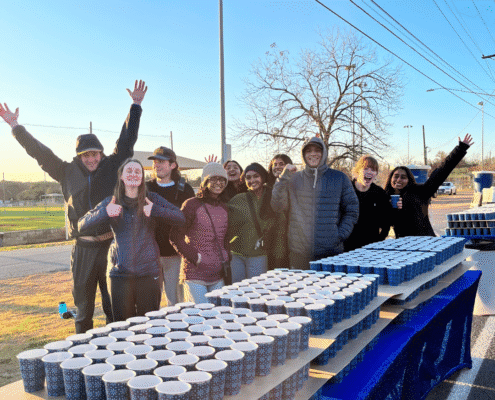 A group of smiling people in jackets stand behind tables lined with rows of paper cups, showcasing volunteer opportunities outdoors on a sunny day, with some raising their arms and a leafless tree and blue sky in the background. Austin Marathon Half Marathon & 5K