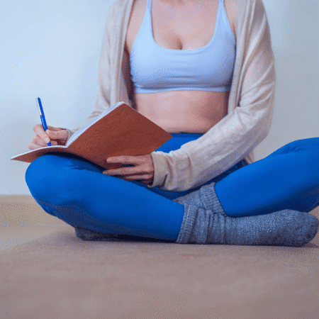 A woman in blue leggings, gray socks, and a sports bra sits cross-legged on a mat, journaling in a notebook with a pen—reflecting on training. Her face is not visible. Austin Marathon Half Marathon & 5K