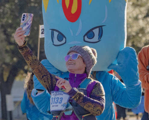 A woman in running gear takes a selfie with a person in a large blue costume featuring yellow flames at an outdoor event highlighting volunteer opportunities. She smiles brightly, wearing sunglasses and a beanie. Austin Marathon Half Marathon & 5K